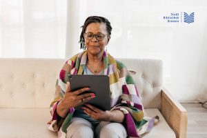 Frau mit Brille sitzt auf einem Sofa und hält ein Tablet in den Händen